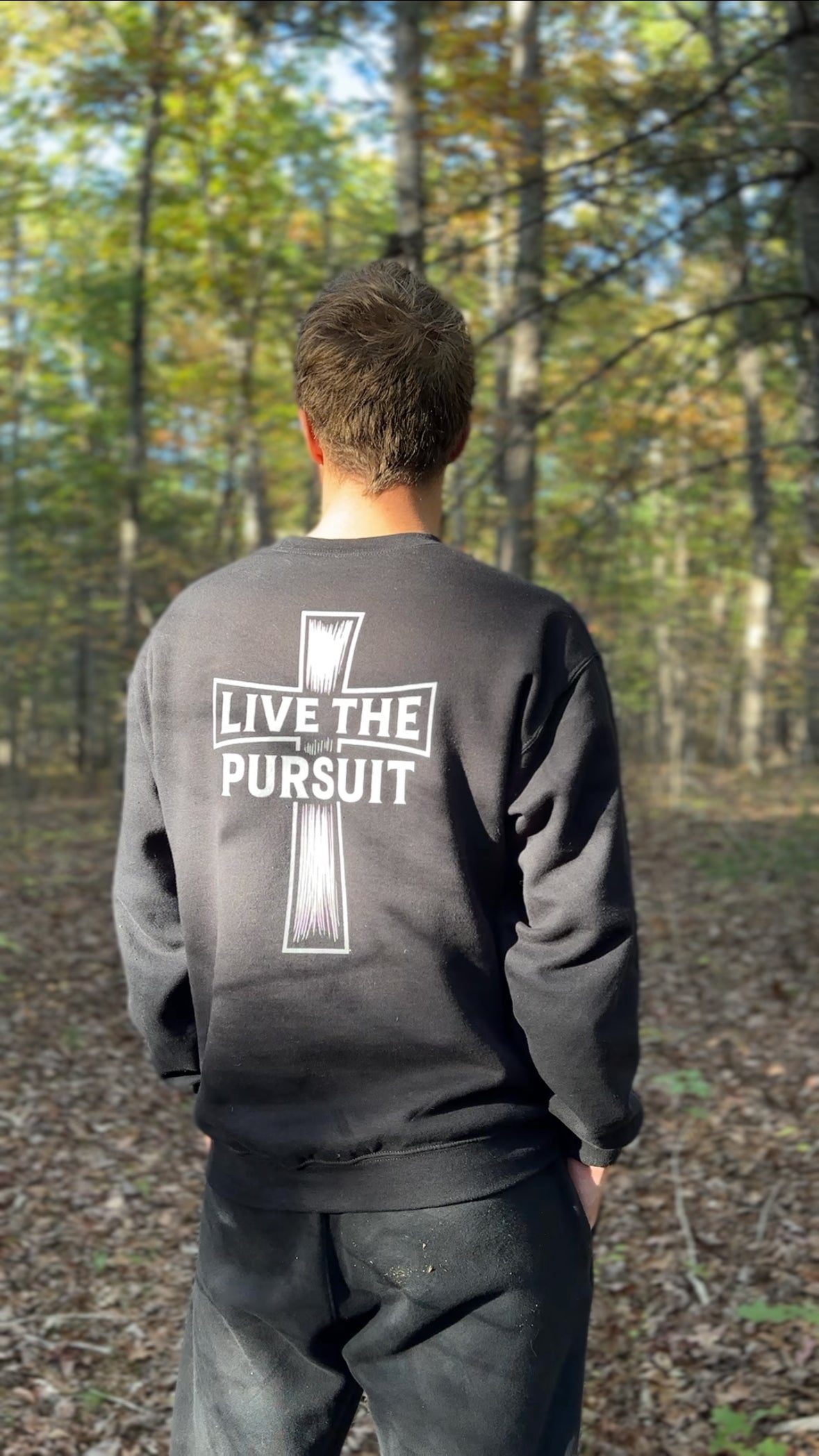 Person wearing the Rack & River Outdoors ‘Live the Pursuit’ cross crewneck, shown from the back while standing in a forest.