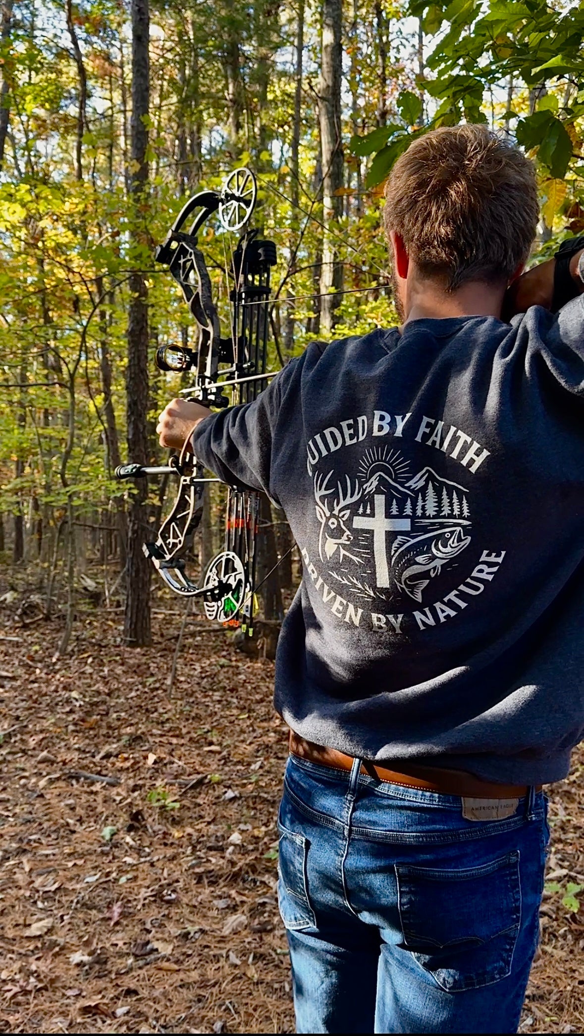 Man drawing a compound bow in the woods while wearing a Rack & River Outdoors sweatshirt with the ‘Guided by Faith, Driven by Nature’ design on the back.