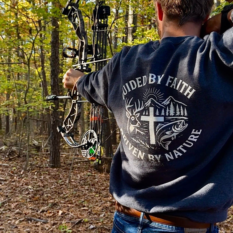 Man drawing a compound bow in the woods while wearing a Rack & River Outdoors sweatshirt with the ‘Guided by Faith, Driven by Nature’ design on the back.
