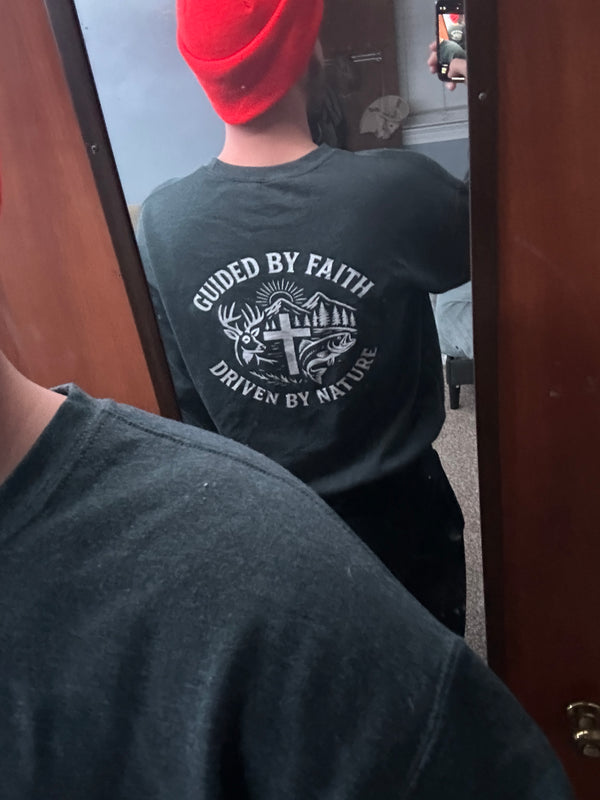 Man wearing a Rack & River Outdoors t-shirt with ‘Guided by Faith, Driven by Nature’ Christian outdoor design, featuring a cross, deer antlers, fish, and mountain scene, photographed in a mirror. Faith-based outdoor apparel for hunters and anglers.
