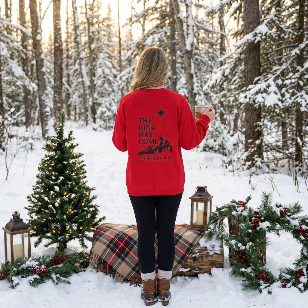Woman wearing the Rack & River Outdoors red ‘The King Has Come’ Christmas sweatshirt, standing in a snowy forest with holiday decor.